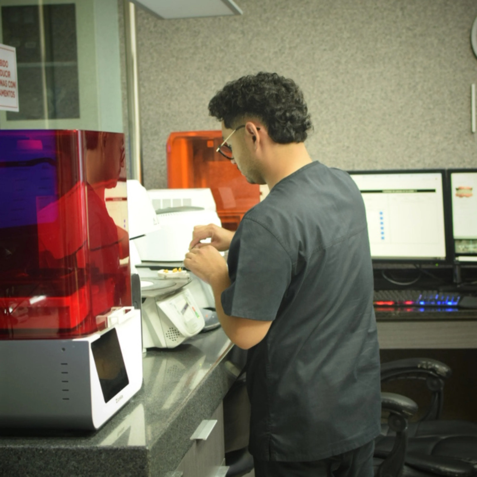 Técnico de Dent Lab manipulando piezas pequeñas sobre una charola junto a las impresoras de resina del laboratorio