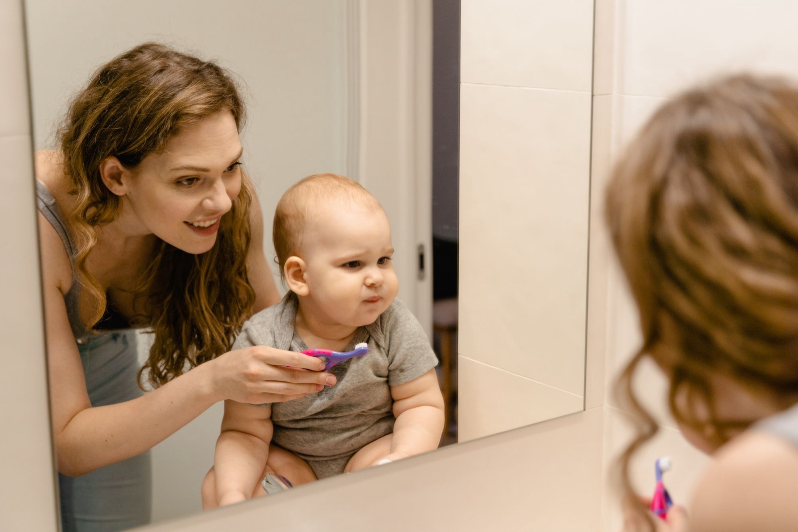 Una mamá acerca un cepillo dental pequeño a su bebé frente al espejo del baño