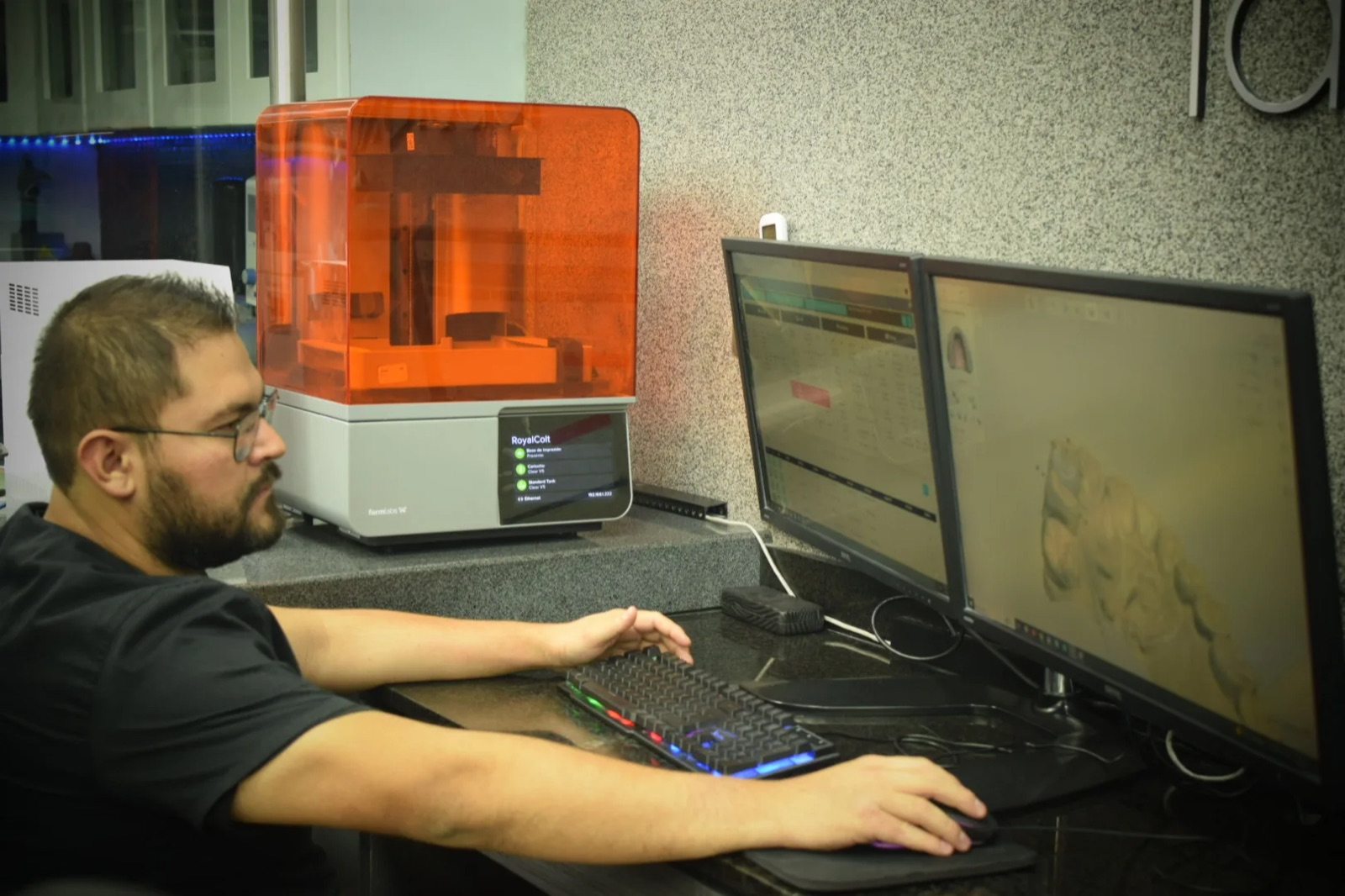 Técnico de laboratorio dental trabaja frente a monitores con modelos dentales digitales, junto a una impresora 3D